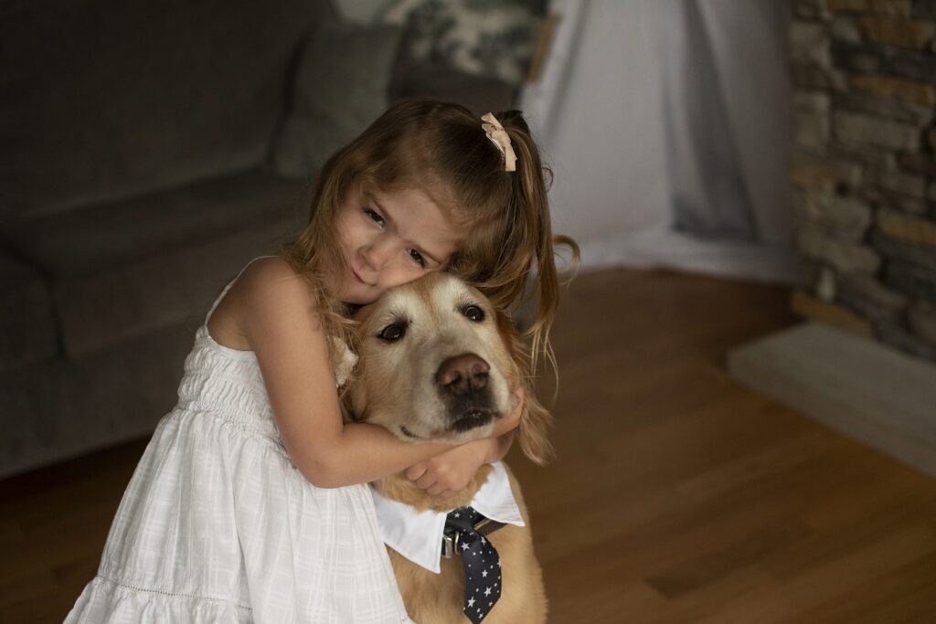 Little girl in white dress with her arms lovingly wrapped around her pet dog who is wearing a tie.