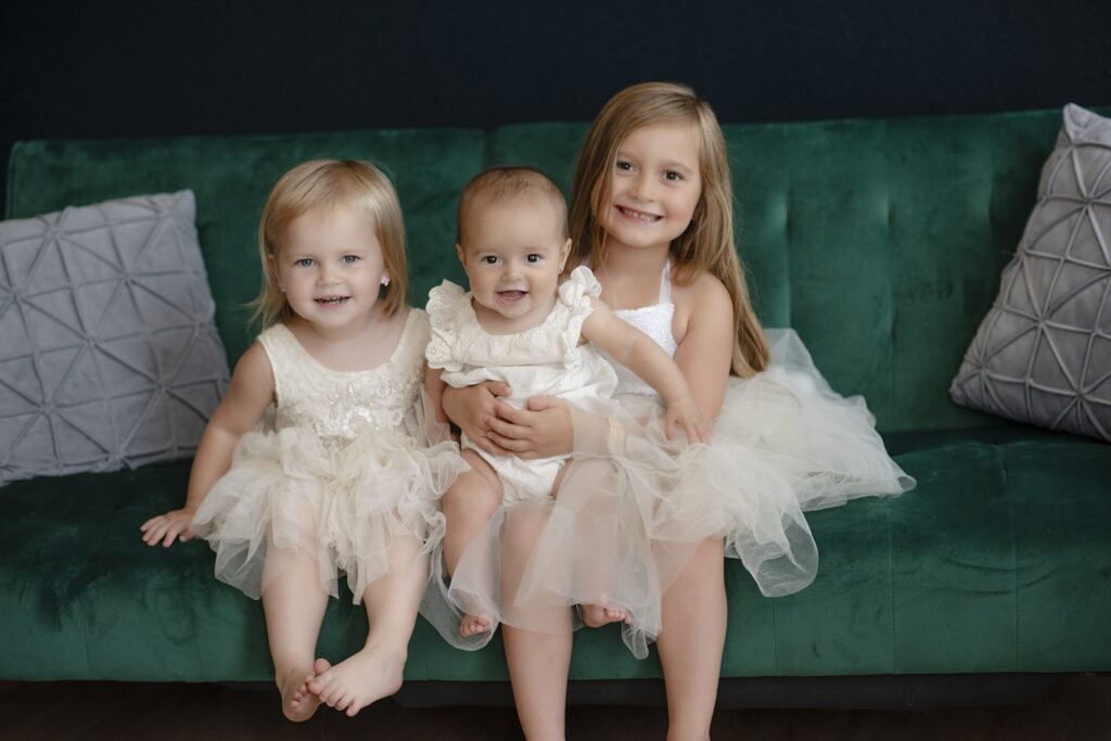 Studio portrait of three little girls wearing white dresses seated on a plush green couch.