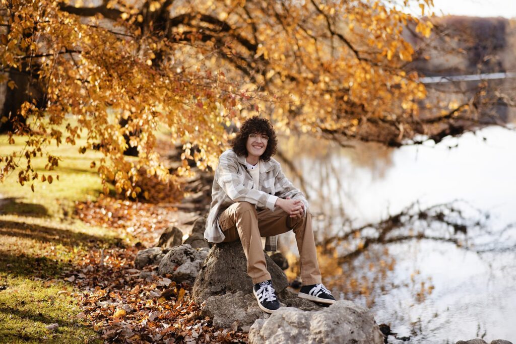 Smiling young man seated on a large rock, surrounded by golden leaves during senior portrait session with Henwood Studios.