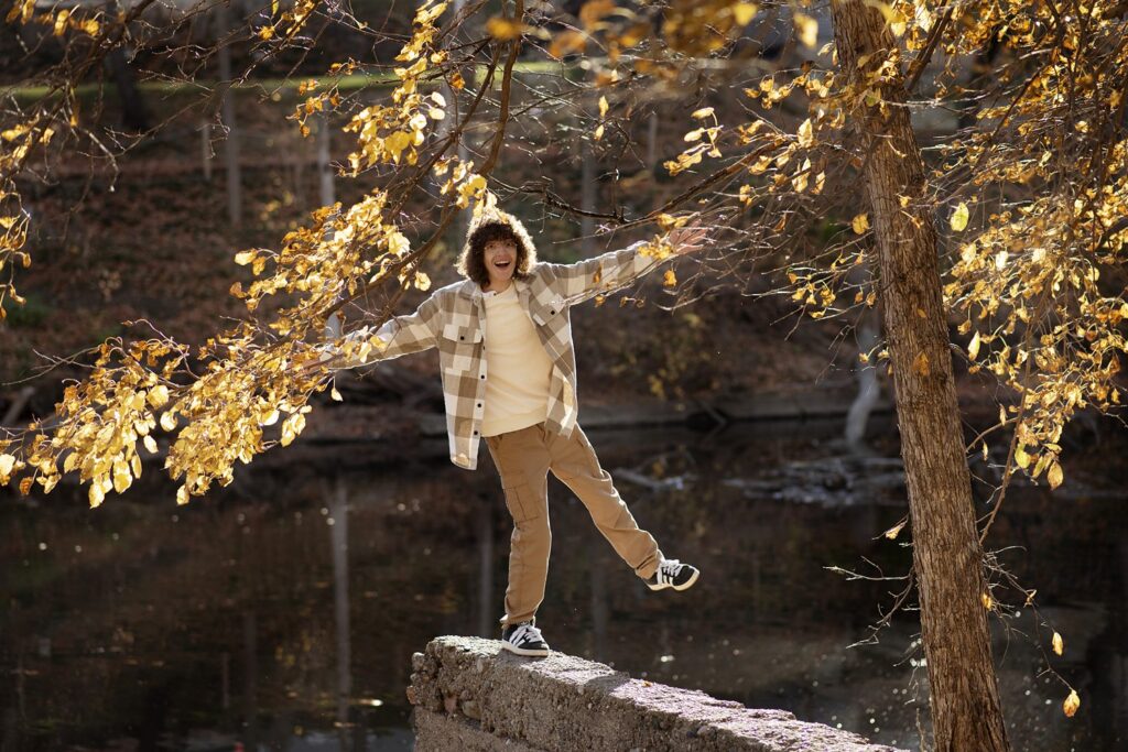 Playful senior photo of young man with arms spread wide balancing on one leg atop a stone wall.