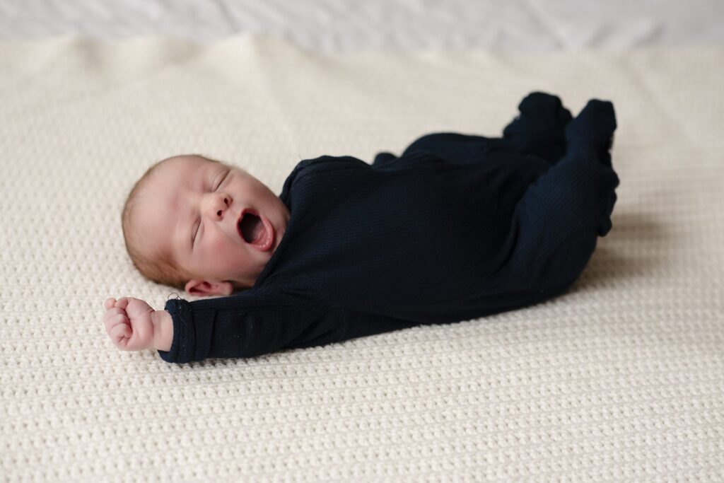 Photo of newborn boy yawning with his arm raised, lying on a cream colored knit blanket.