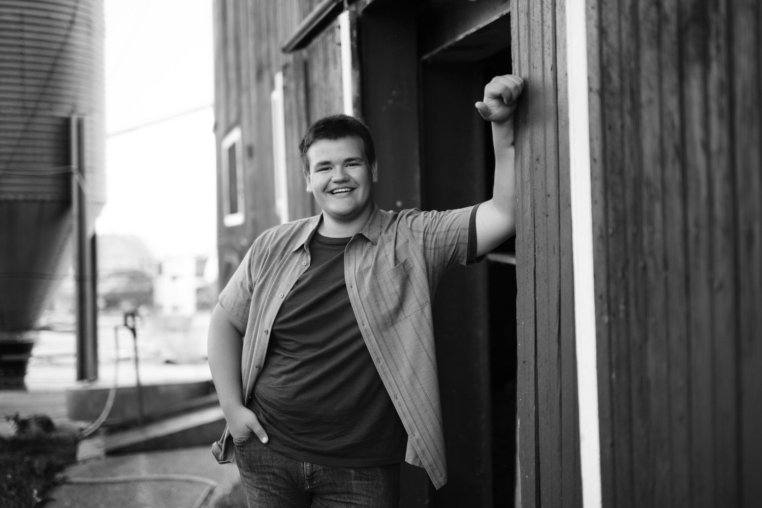 Black and white photo of high school senior smiling, leaning against barn.