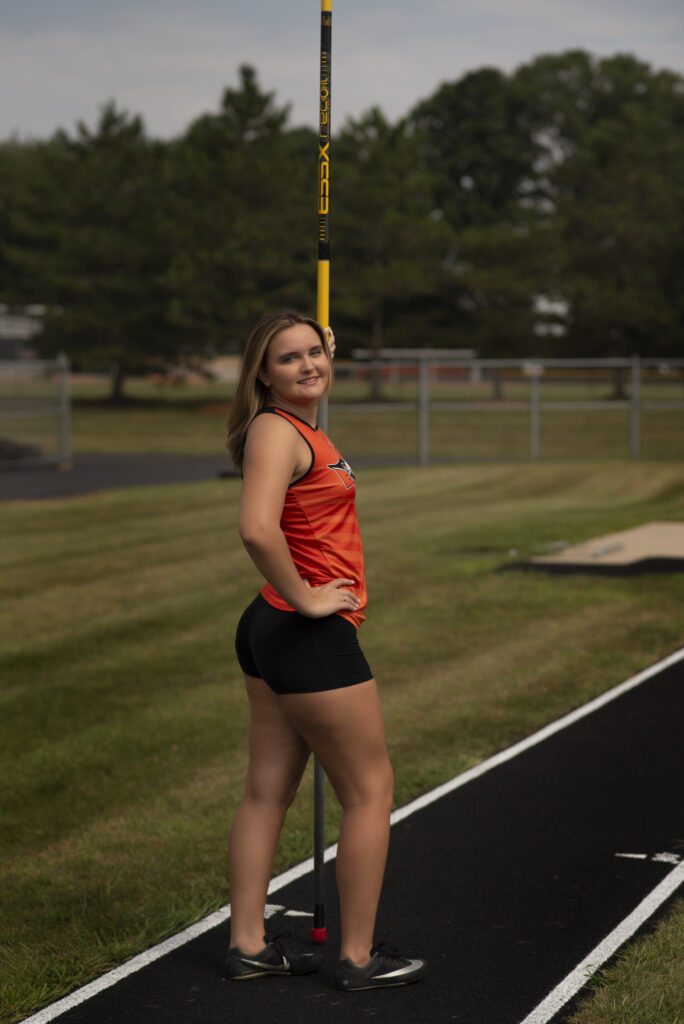 Track athlete standing in uniform with her javelin during senior photo session with Henwood Studios. 