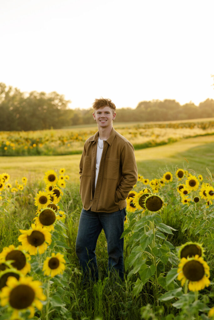 Smiling high school senior standing a a row of sunflowers during session with Henwood Studios.
