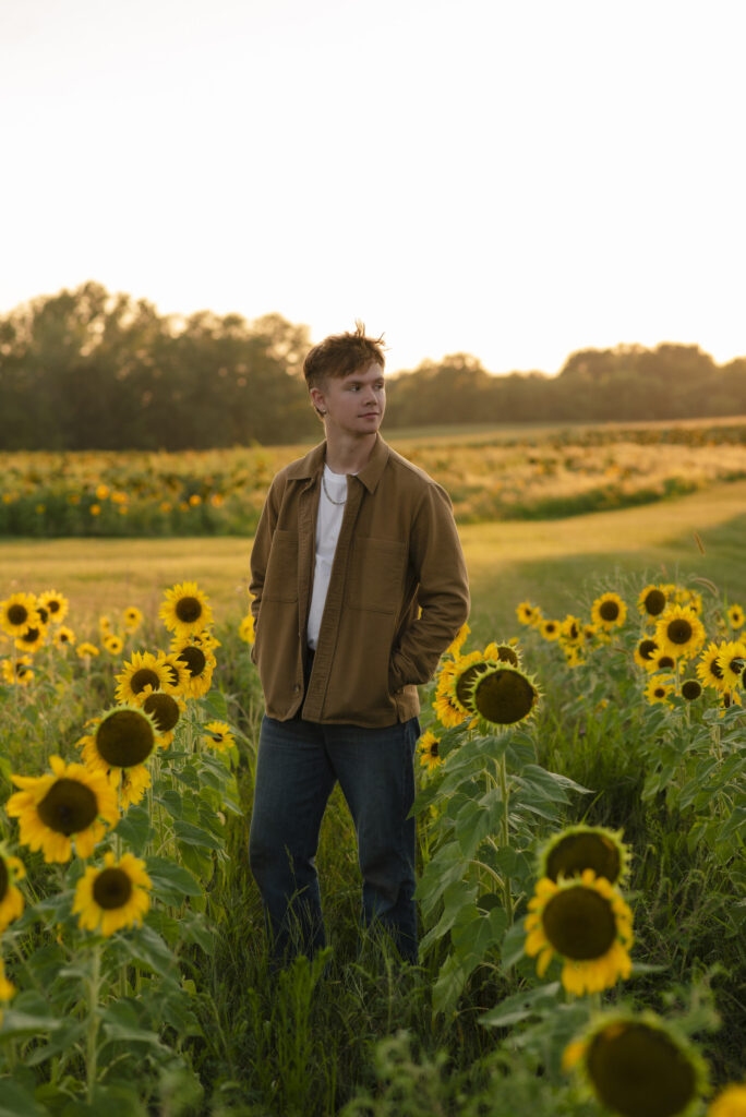 Senior portrait of young man wearing a brown jacket walking through a sunflower field.