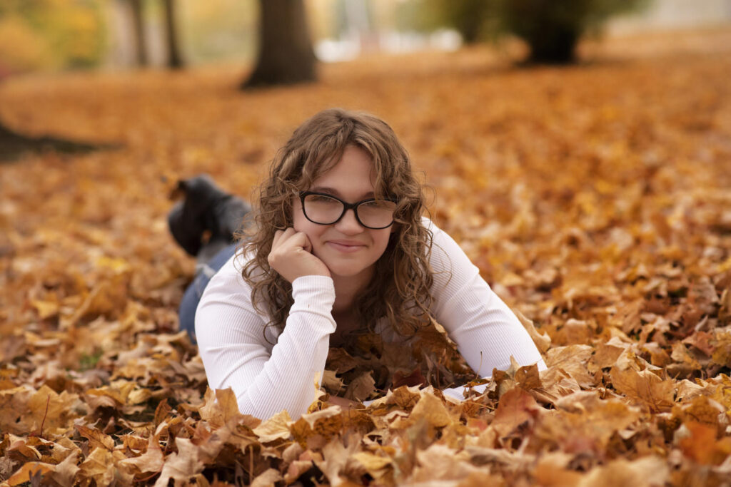 Solo portrait of young girl in white sweater lying on orange leaves.