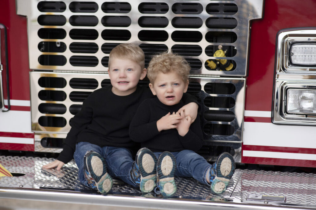 Two little brothers with their arms around each other looking at the camera, sitting on a fire truck.