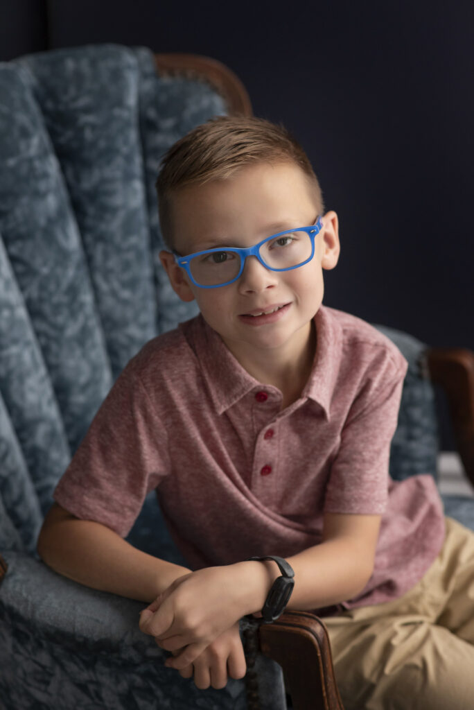 Portrait of young boy wearing glasses seated on plush chair.