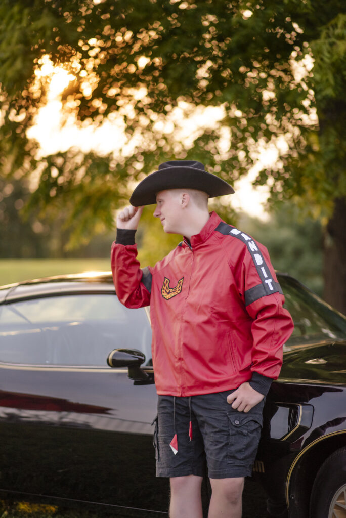 High school senior wearing a red jacket, tipping his cowboy hat leaning against Pontiac Trans Am.